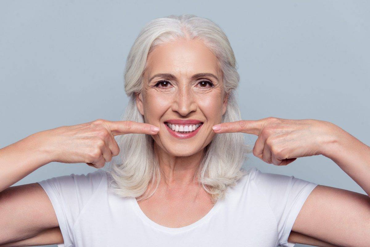 Concept of having strong healthy straight white teeth at old age. Close up portrait of happy with beaming smile female pensioner pointing on her perfect clear white teeth, isolated on gray background